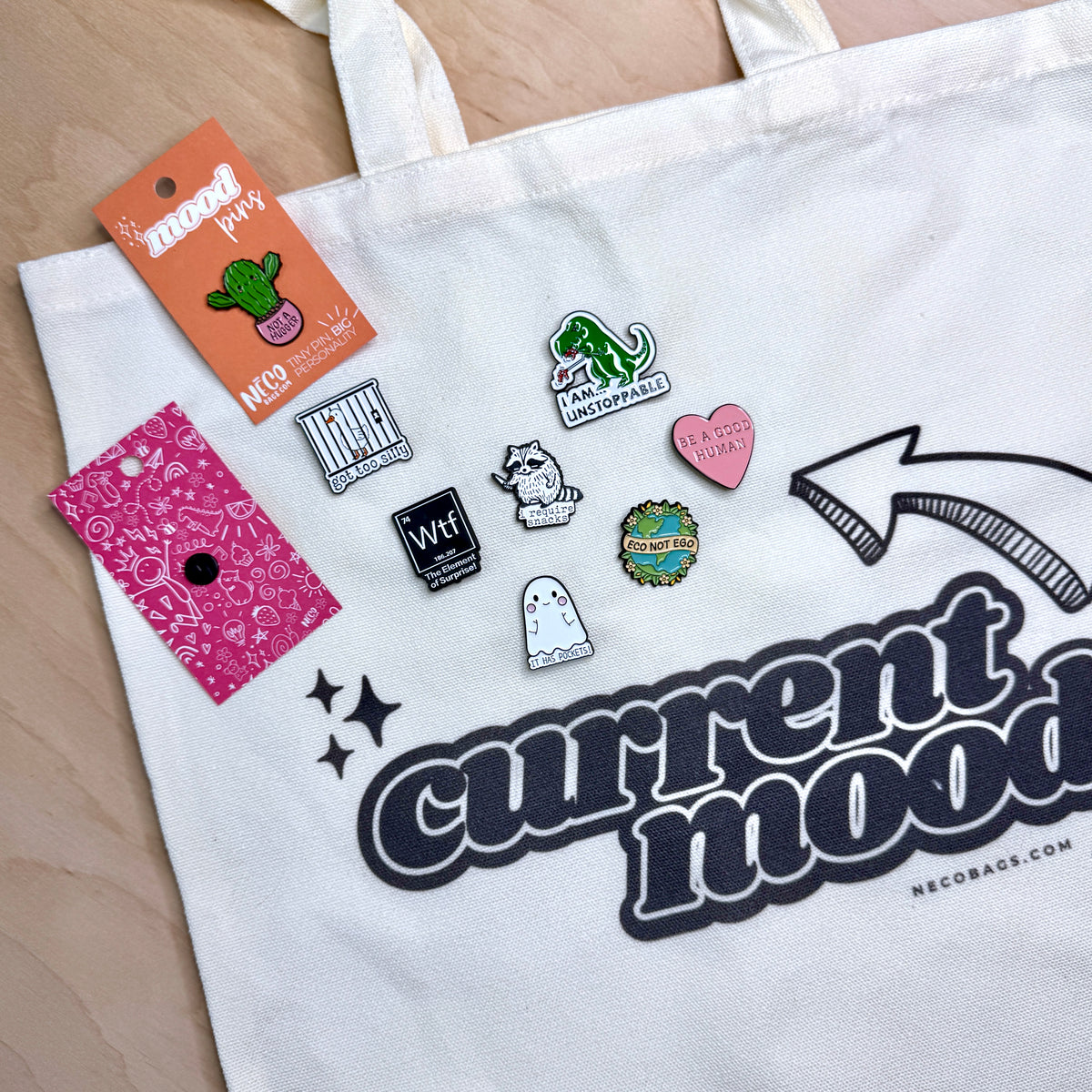 A white canvas tote bag with the words 'current mood' printed in large black letters, shown on a light wood surface. Several enamel pins are clipped onto the bag, including a green dinosaur pin, a pink heart pin, a ghost pin, and a cactus tag labeled 'mood pins' and 'NECOBAGS'.