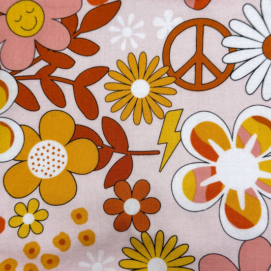 A close-up of a retro 1960s and 1970s style fabric print featuring yellow, orange, and pink flowers, brown leaves, a peace sign, a lightning bolt, and daisies on a dusty pink background.