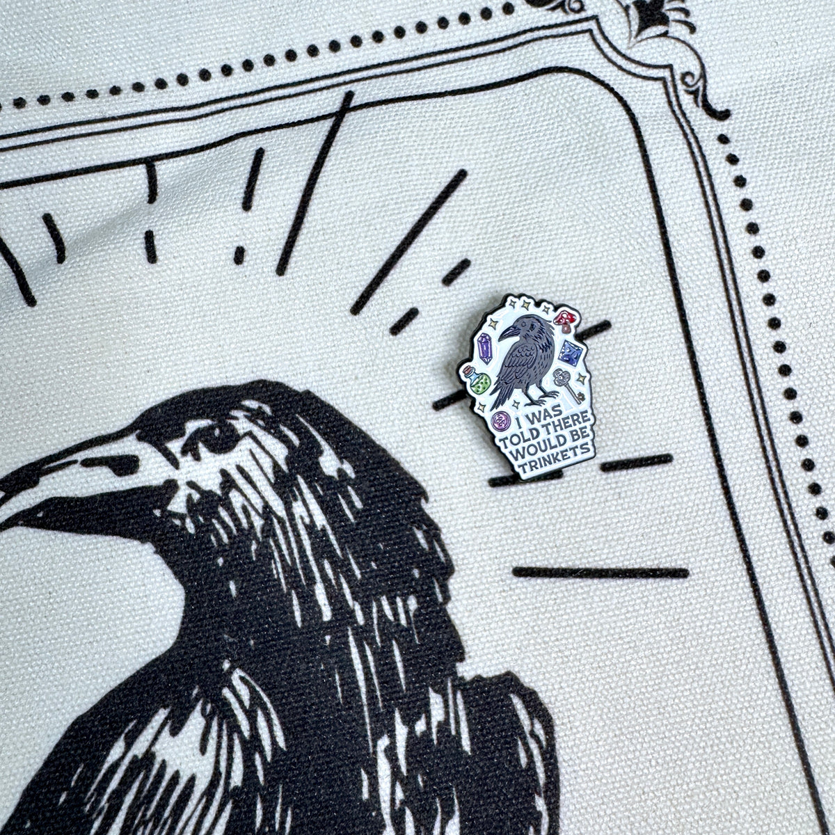 A small enamel pin shaped like a white cloud with a raven illustration and the text 'I WAS TOLD THERE WOULD BE TRINKETS' is shown pinned to a white fabric with a black raven graphic.