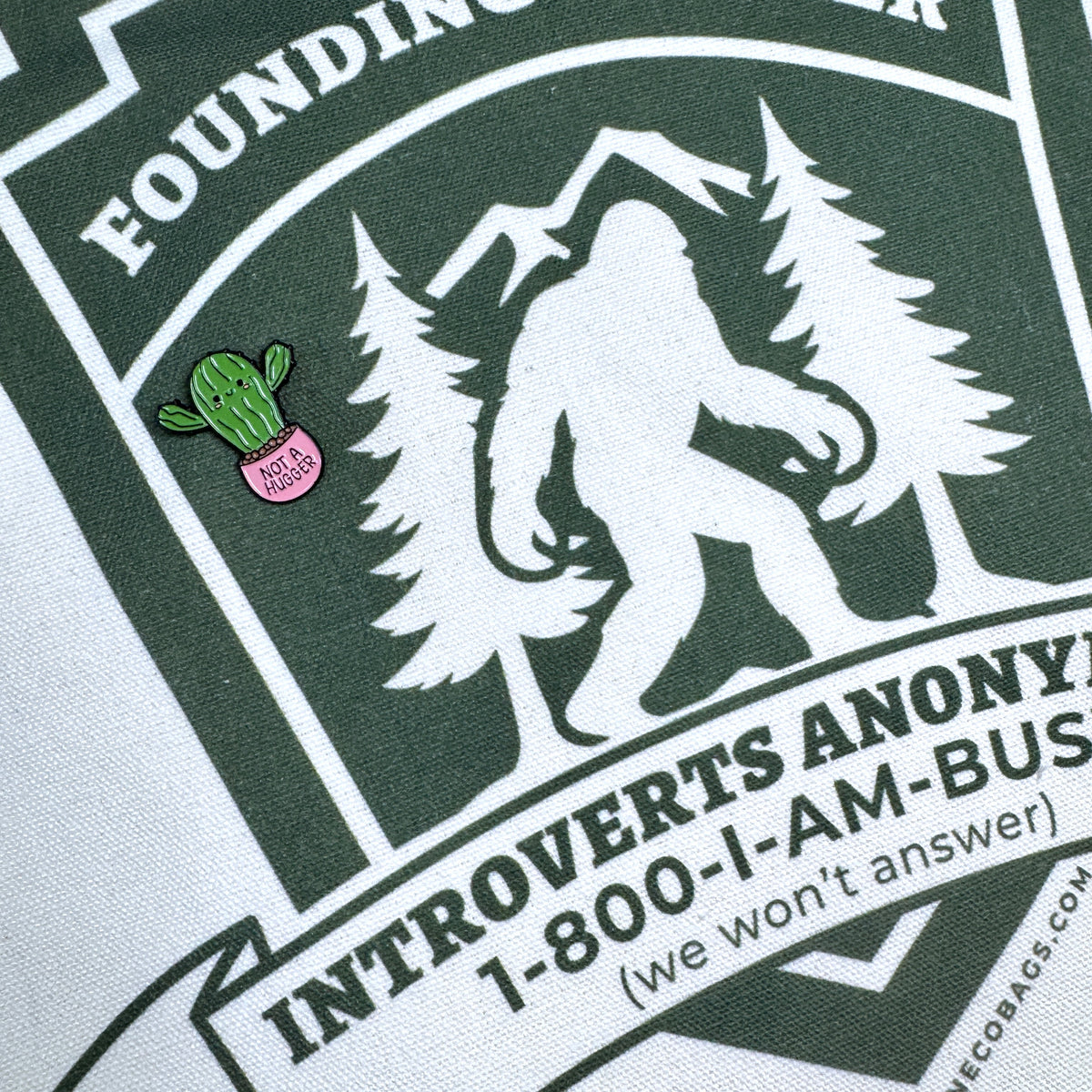 A close-up of a dark green and white printed graphic on fabric, featuring a Bigfoot silhouette between trees with the text 'Founding Members' and 'Introverts Anonymous. A small pink cactus enamel pin is pinned to the left side of the print.