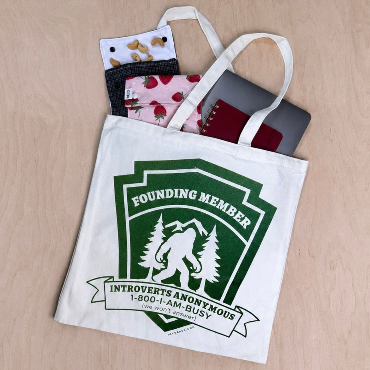 A white canvas tote bag with a green and white 'Founding Member' graphic, shown filled with various patterned fabric items on a light wood background.