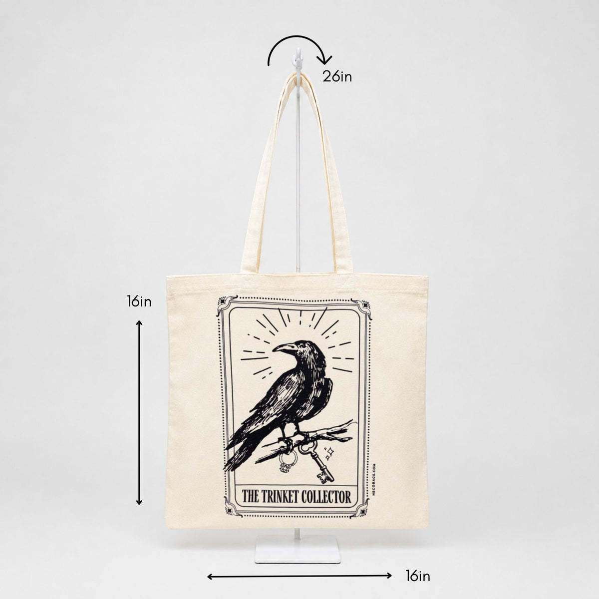 An off-white canvas tote bag with a black raven graphic and the text 'THE TRINITY COLLECTOR' on the front, shown hanging on a white display stand against a light gray background with dimension arrows indicating 16in height and 16in width, plus a 26in handle length.