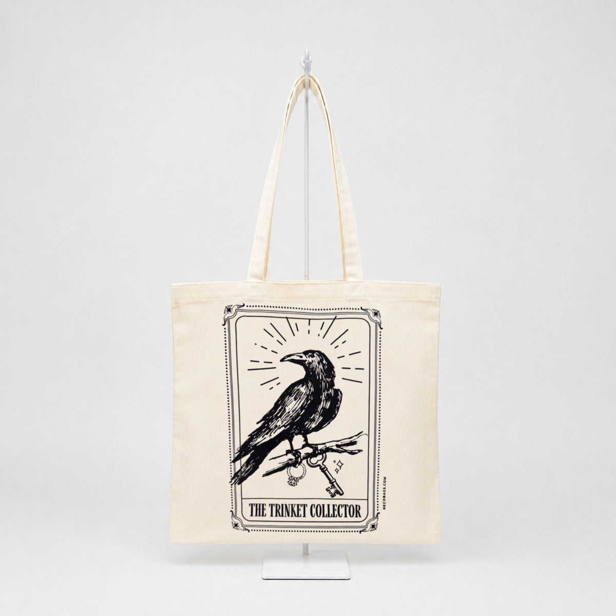 An off-white canvas tote bag with long handles, featuring a black graphic print of a raven perched on a branch inside a bordered frame, with the text 'THE TRINKET COLLECTOR' below. The bag is displayed on a white stand against a light gray background.