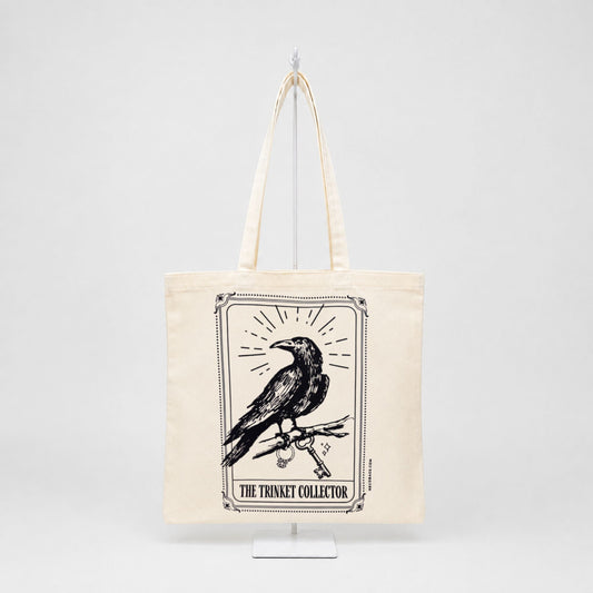 An off-white canvas tote bag with long handles, featuring a black graphic print of a raven perched on a branch inside a bordered frame, with the text 'THE TRINKET COLLECTOR' below. The bag is displayed on a white stand against a light gray background.