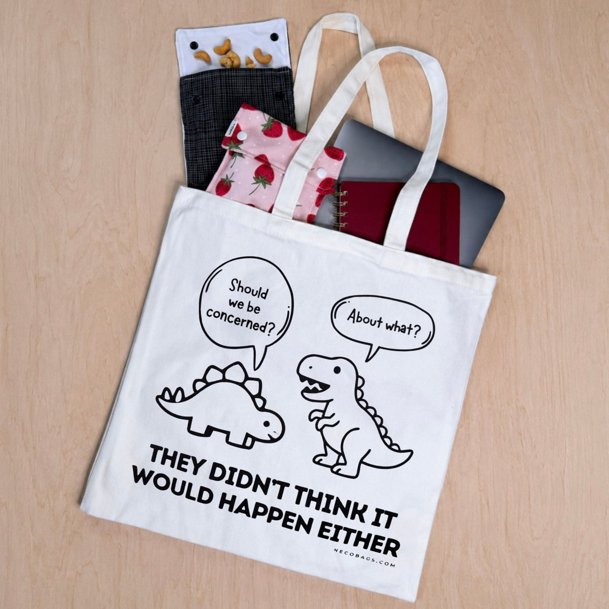 A white canvas tote bag with a black cartoon dinosaur graphic and the text 'THEY DIDN'T THINK IT WOULD HAPPEN EITHER' lies on a light wood surface. Partially tucked inside the bag are patterned snack bags notebook and laptop
