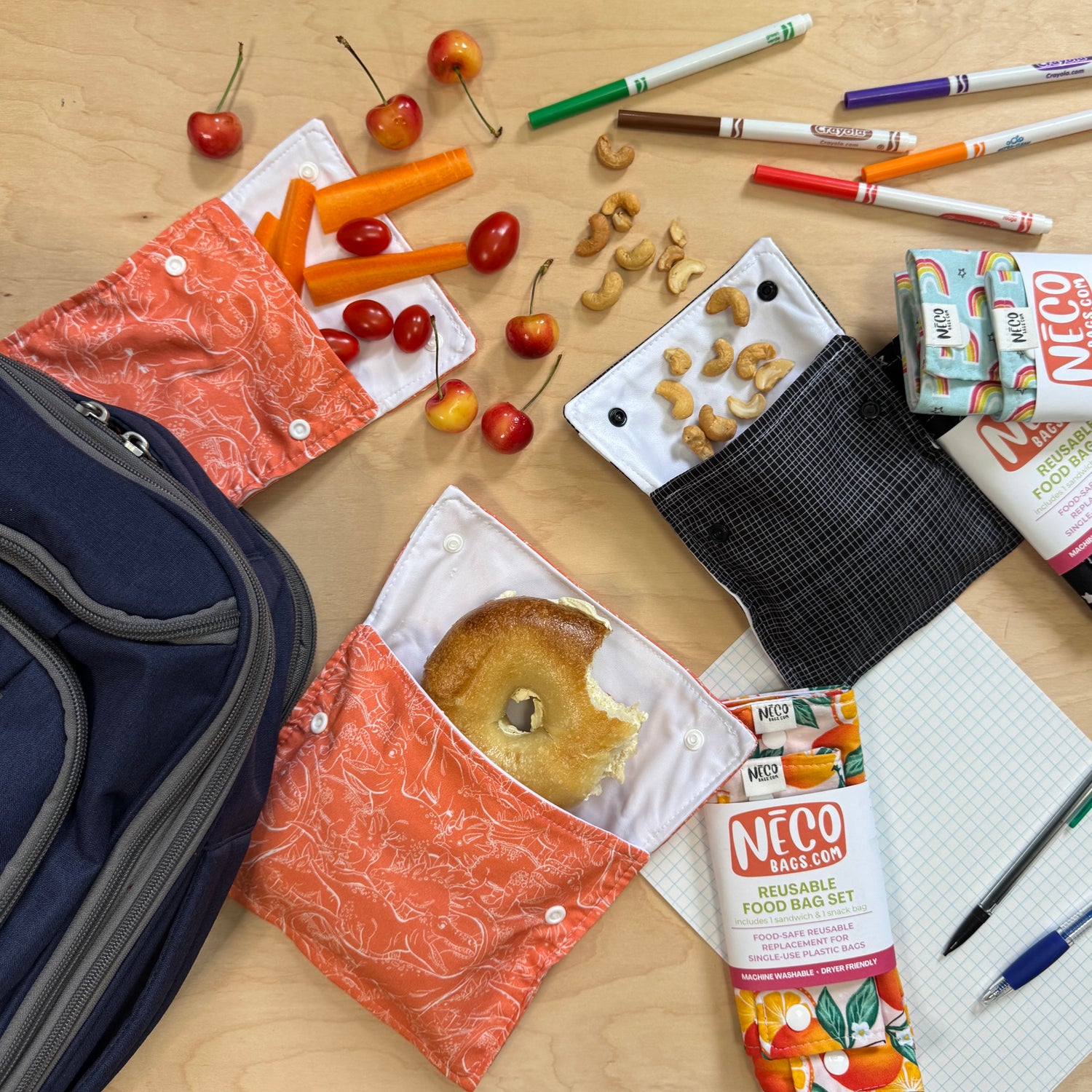 A styled flat-lay of reusable machine washable fabric food bag sets in coral orange and black, shown with school lunch items including cherries, carrots, cashews, a bagel half-eaten, NECO Bags, markers, and a notebook on a light wood surface.