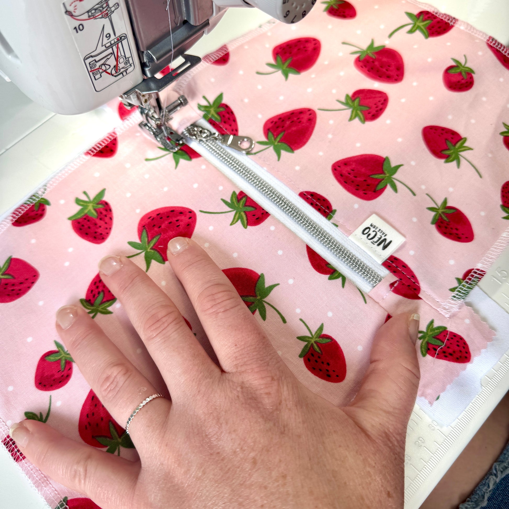 A close-up of a sewing machine stitching a pink fabric with a red strawberry print. A hand is guiding the fabric under the sewing press, Machine washable cosmetic bags