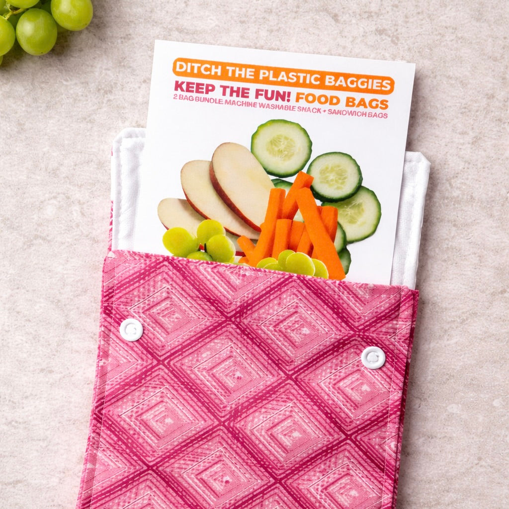 A pink patterned reusable snack bag with snap closures, shown holding a white paper food bag with an info card with sliced vegetables and the text 'Ditch the Plastic Baggies Keep the Fun! Food Bags.' The scene is styled on a light stone surface with grapes partially visible in the corner.