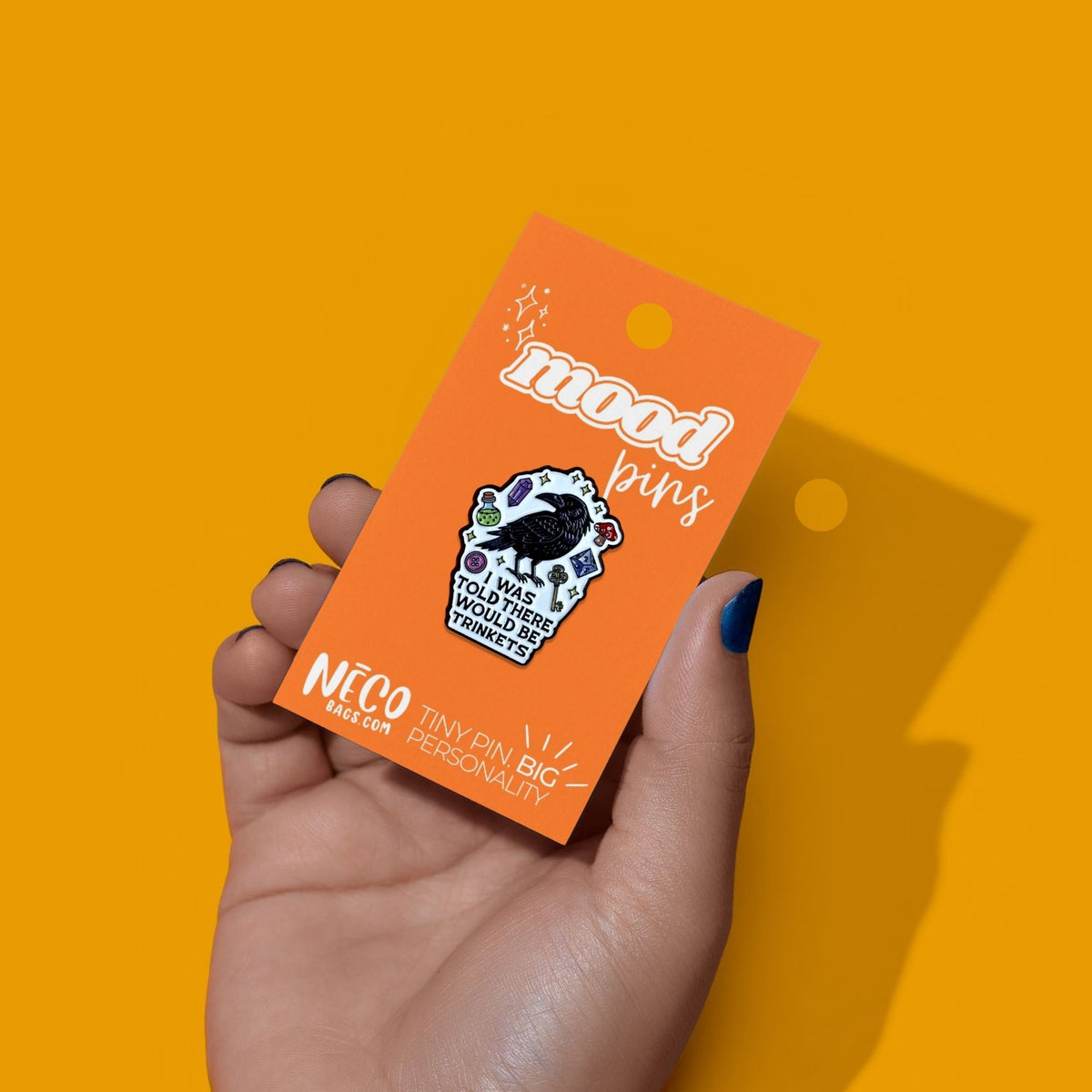 A hand with dark nail polish holds an orange backing card displaying a colorful enamel pin shaped like a cupcake with a black cat on top. The card reads 'mood pins' and 'NICO BIG CAT TINY PIN BIG PERSONALITY'.