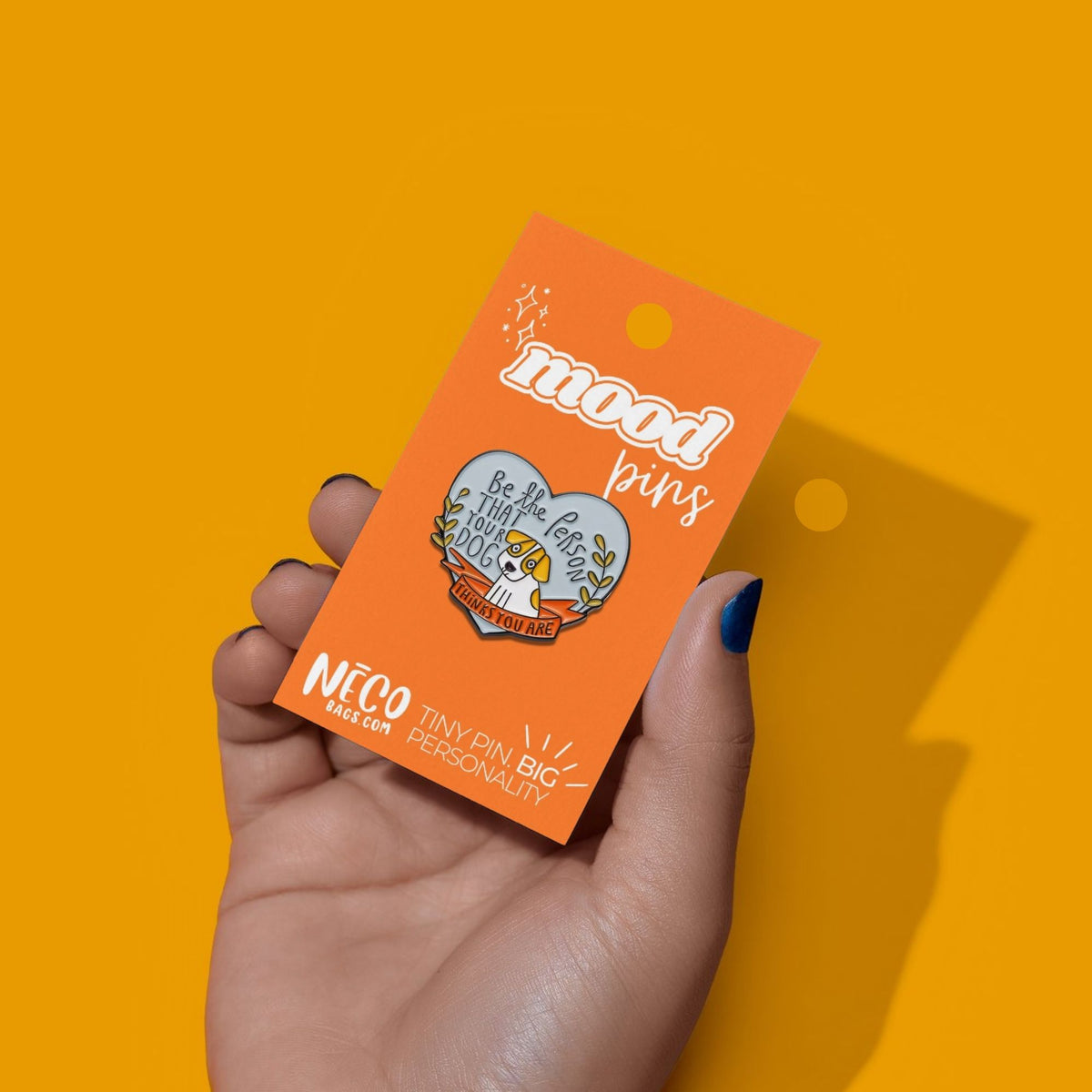 A hand with dark nail polish holds an orange backing card displaying a heart-shaped enamel pin. The backing card reads 'mood pins' and 'NÉCO BIG CAT'.