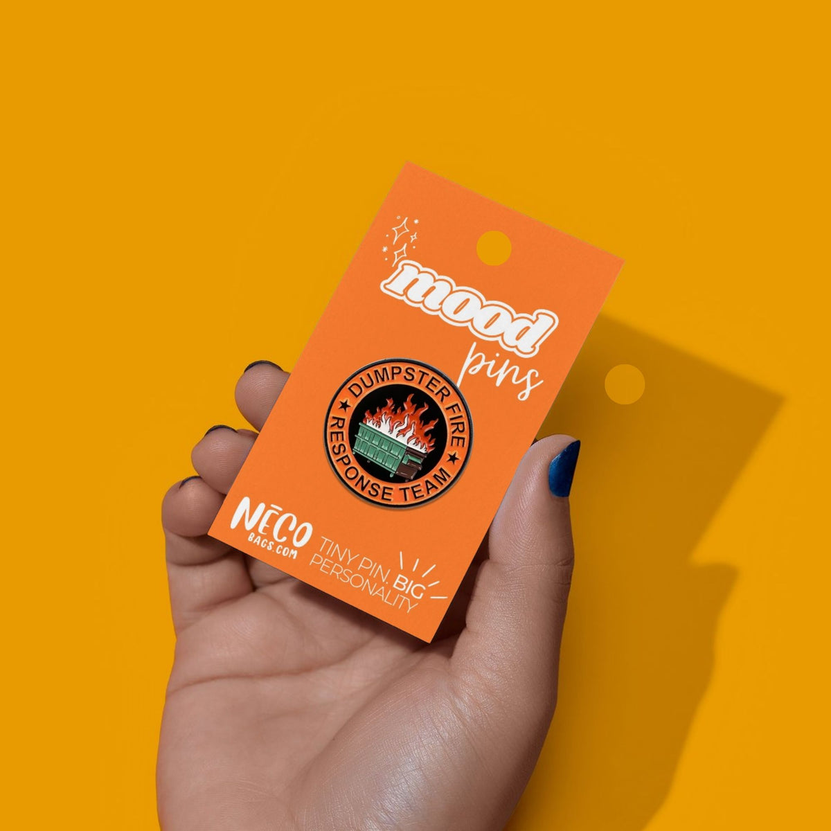 A hand with blue nail polish holds an orange card displaying a 'mood pins' brand 'Humperster Fire Response Team' enamel pin against a solid yellow background.