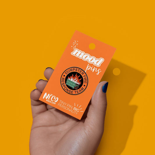 A hand with blue nail polish holds an orange card displaying a 'mood pins' brand 'Humperster Fire Response Team' enamel pin against a solid yellow background.