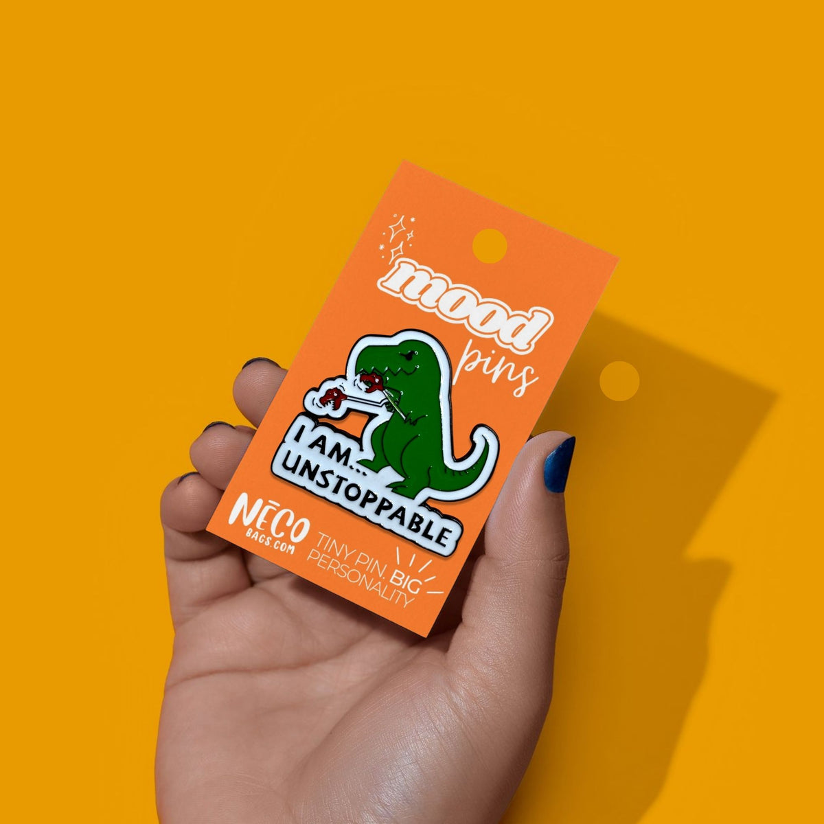 A hand with blue nail polish holds an orange backing card displaying a green dinosaur enamel pin that reads 'I AM UNSTOPPABLE'. The background is a solid, vibrant yellow.