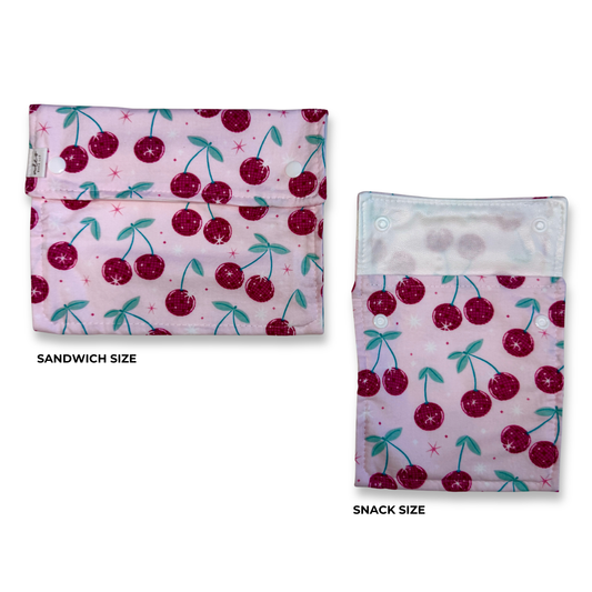 Two fabric snack and sandwich pouches with a pink cherry print are shown on a white background, labeled 'SANDWICH SIZE' and 'SNACK SIZE'. the cherries are a fun bright colorful pink disco balls. Machine washable food safe bags