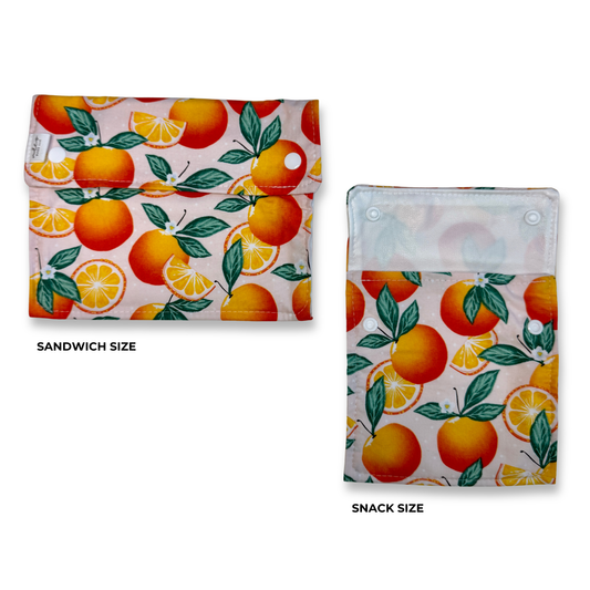 Two fabric snack pouches with an orange and citrus slice print on a light pink background, shown side by side labeled 'SANDWICH SIZE' and 'SNACK SIZE'. The pouches have a light pink background with bright colorful oranges.  Cute fun, and colorful machine washable food bags