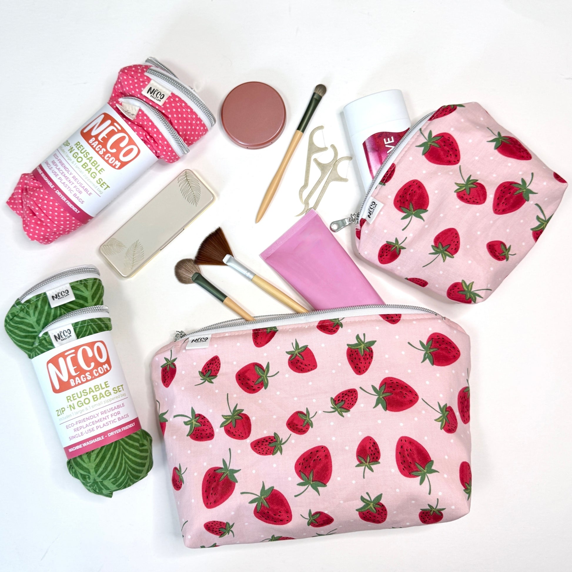 A styled flat-lay of two pink zippered pouches with a strawberry print, surrounded by various colorful knitting and crafting supplies on a clean white background. Machine washable cosmetic bags