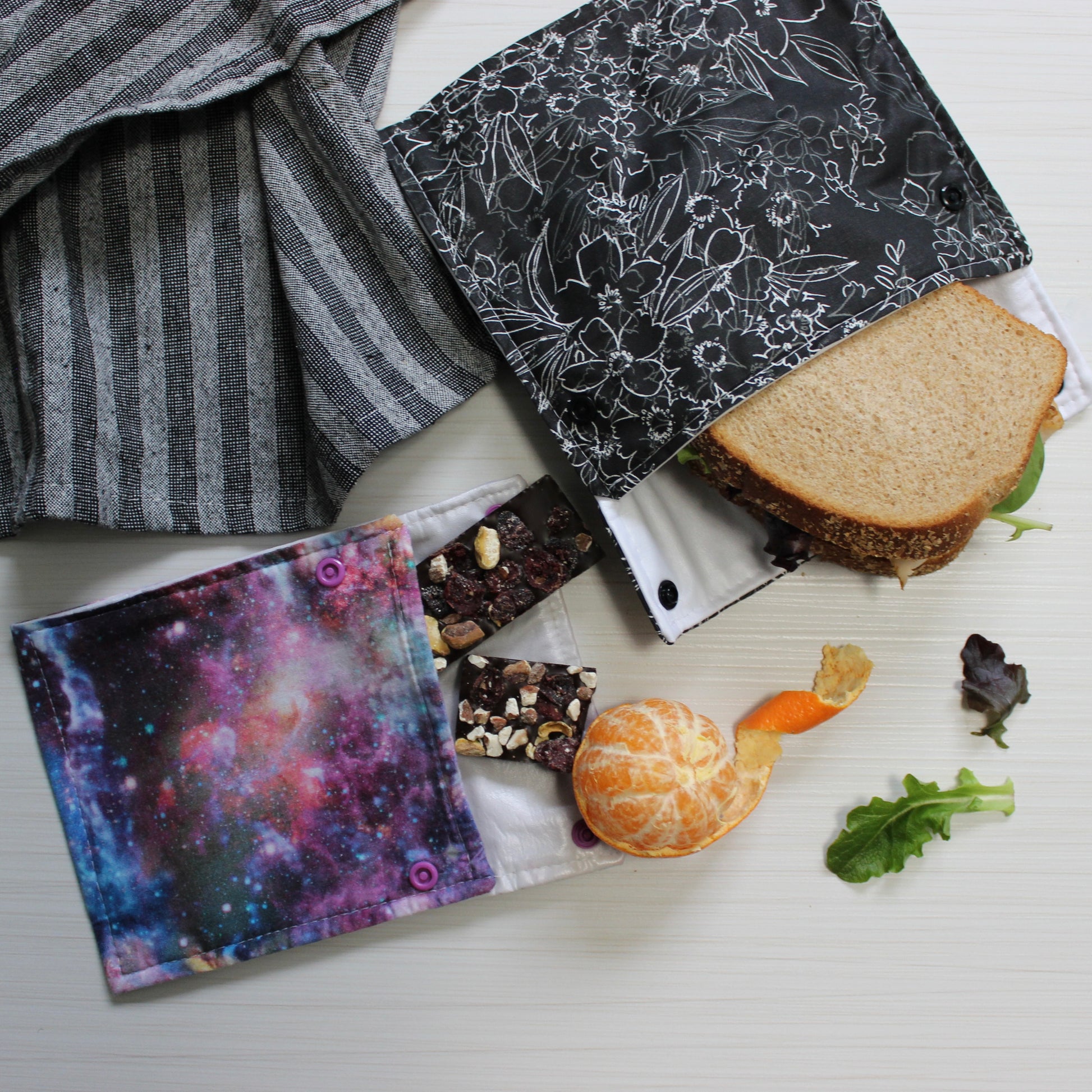 Reusable food bags with different prints next to a sandwich and snacks on a white surface. Snack and Sandwich Bags