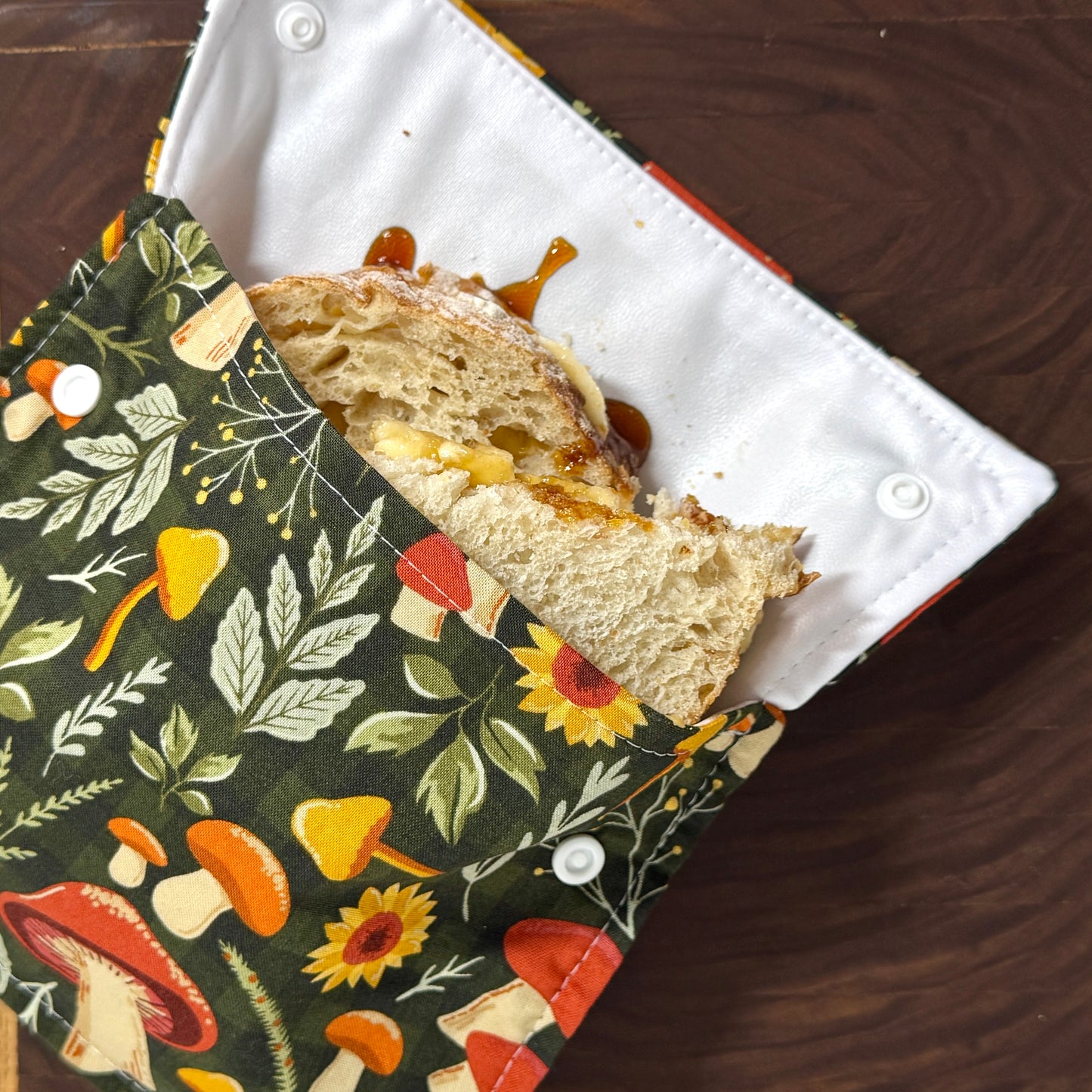 A close-up of a fabric sandwich bag with a floral mushroom print, shown with a large sandwich inside on a dark wooden table. Machine washable sandwich bags and snack bags