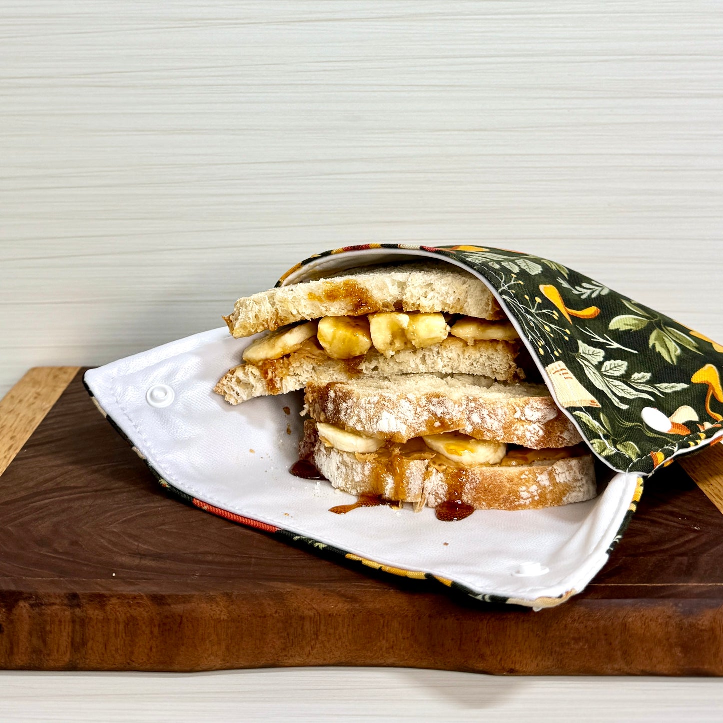 A close-up of a fabric sandwich bag with a floral mushroom print, shown with a large sandwich inside on a dark wooden table. Machine washable snack bags and sandwich bags