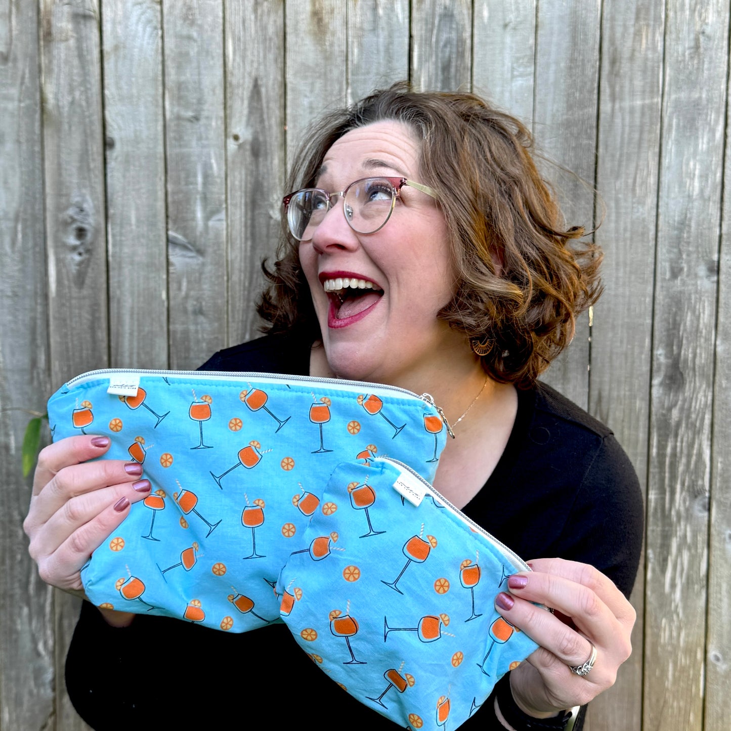 A woman with glasses and brown hair is smiling excitedly while holding a light blue zippered pouch decorated with an orange and black cocktail glass pattern, set against a wooden fence background. Machine washable cosmetic bags